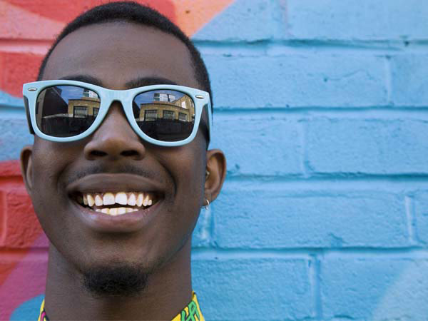 Close up of smiling young man’s face who is wearing blue framed sunglasses while standing in front of a colorfully-painted brick wall
