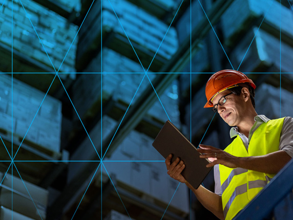 Warehouse worker wearing a hard hat and safety vest using a tablet in front of storage racks.