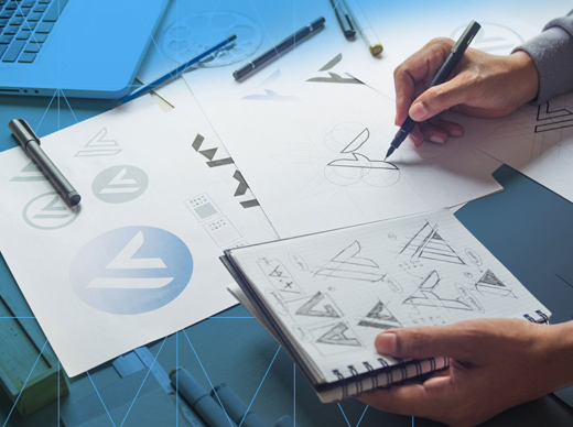 Person sketching logo concepts on paper with design tools spread across a desk.