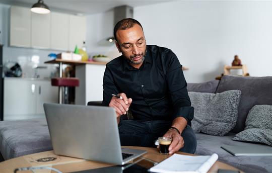 Middle-aged entrepreneur managing his business from home, sitting in front of laptop, reaching for espresso