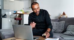 Middle-aged entrepreneur managing his business from home, sitting in front of laptop, reaching for espresso