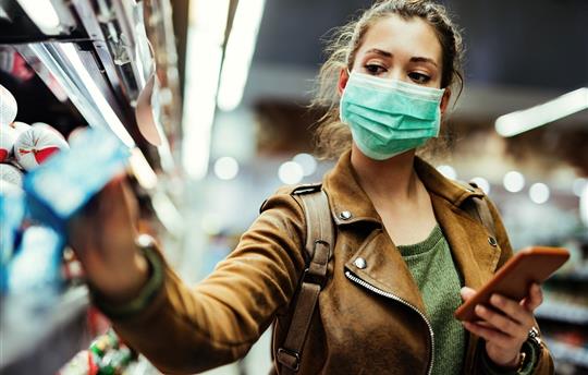 female grocery shopper in brown leather jacket wearing a mask reaching for an item on the shelf, holding smartphone in other hand