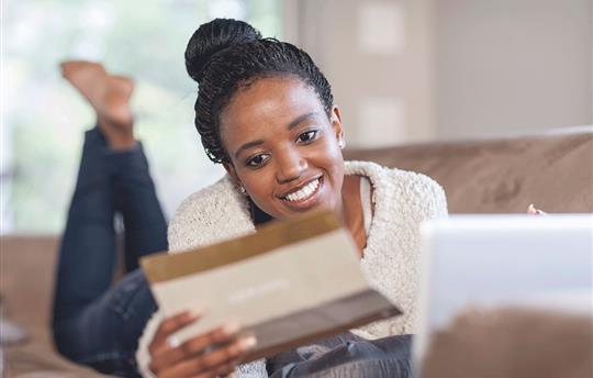 young woman with a smile on her face laying on her couch in front of her laptop holding a small brochure in her hand